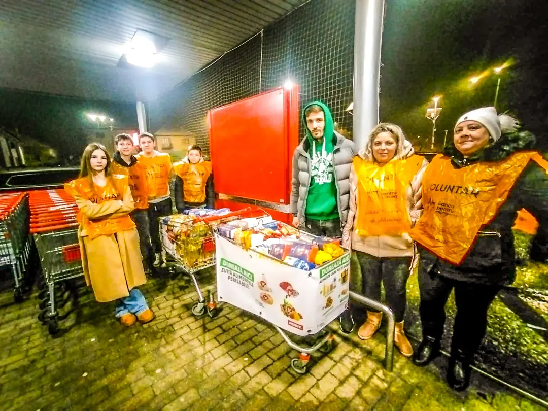 Voluntari cu veste portocalii lângă un coș plin de alimente