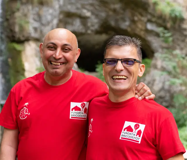 Doi bărbați în tricouri roșii râd, lângă o cascadă